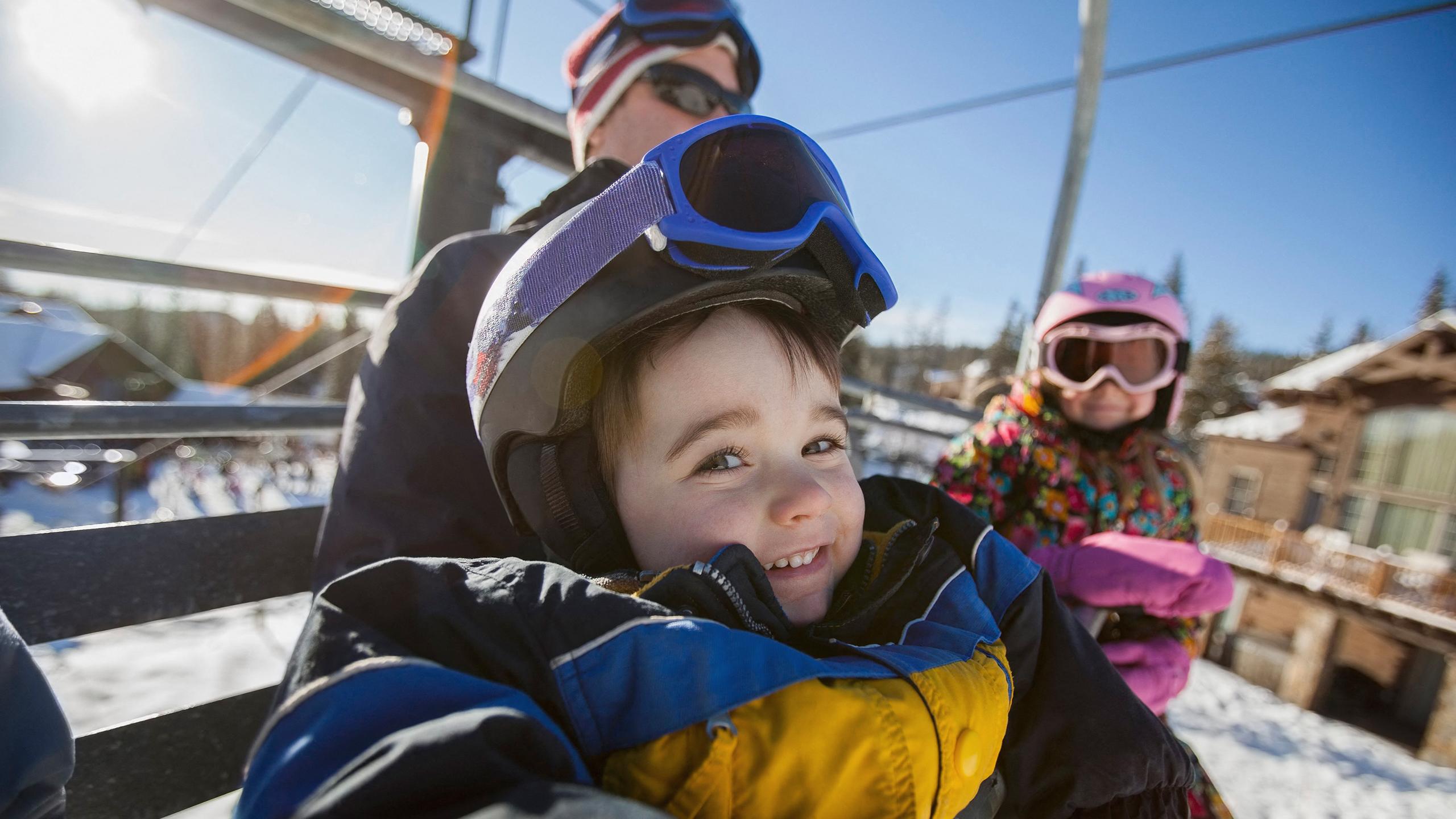 Un enfant est assis sur un télésiège, il sourit emmitouflé dans sa veste de ski - Investir à la montagne - Destination Savoie et Haute-Savoie