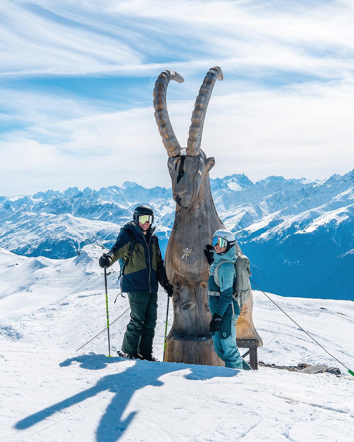 Un couple de personnes posent devant une statut de bouquetin en bois sur les pistes des 3 Vallées